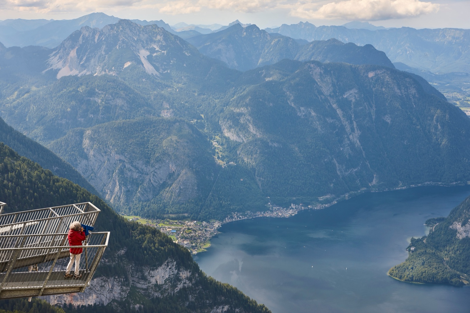 A visitor in a red jacket on a metal viewing platform high above the lake and a village at the foot of forested mountains