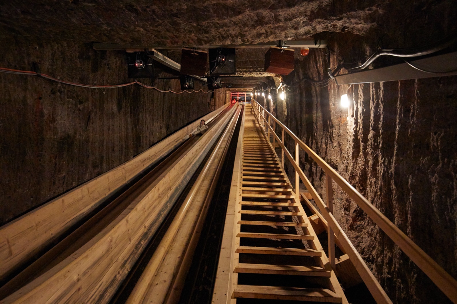 A long wooden slide beside a staircase, running down a lit tunnel cut through rock