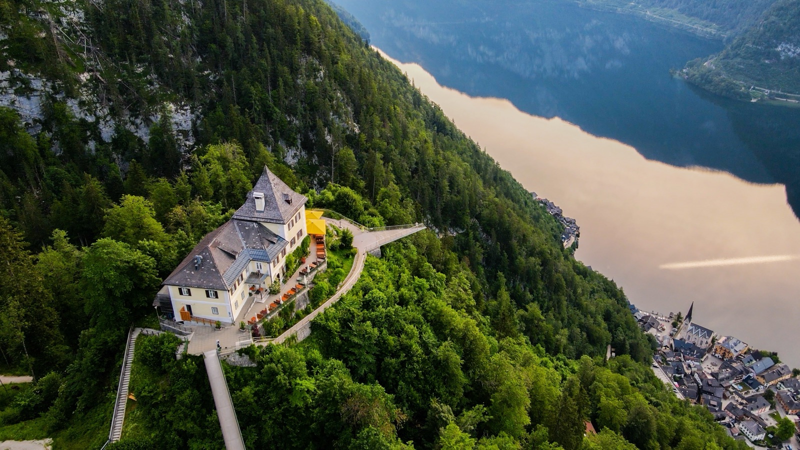 Aerial view of a building and walkway on a wooded spur high above the lake and the rooftops of Hallstatt